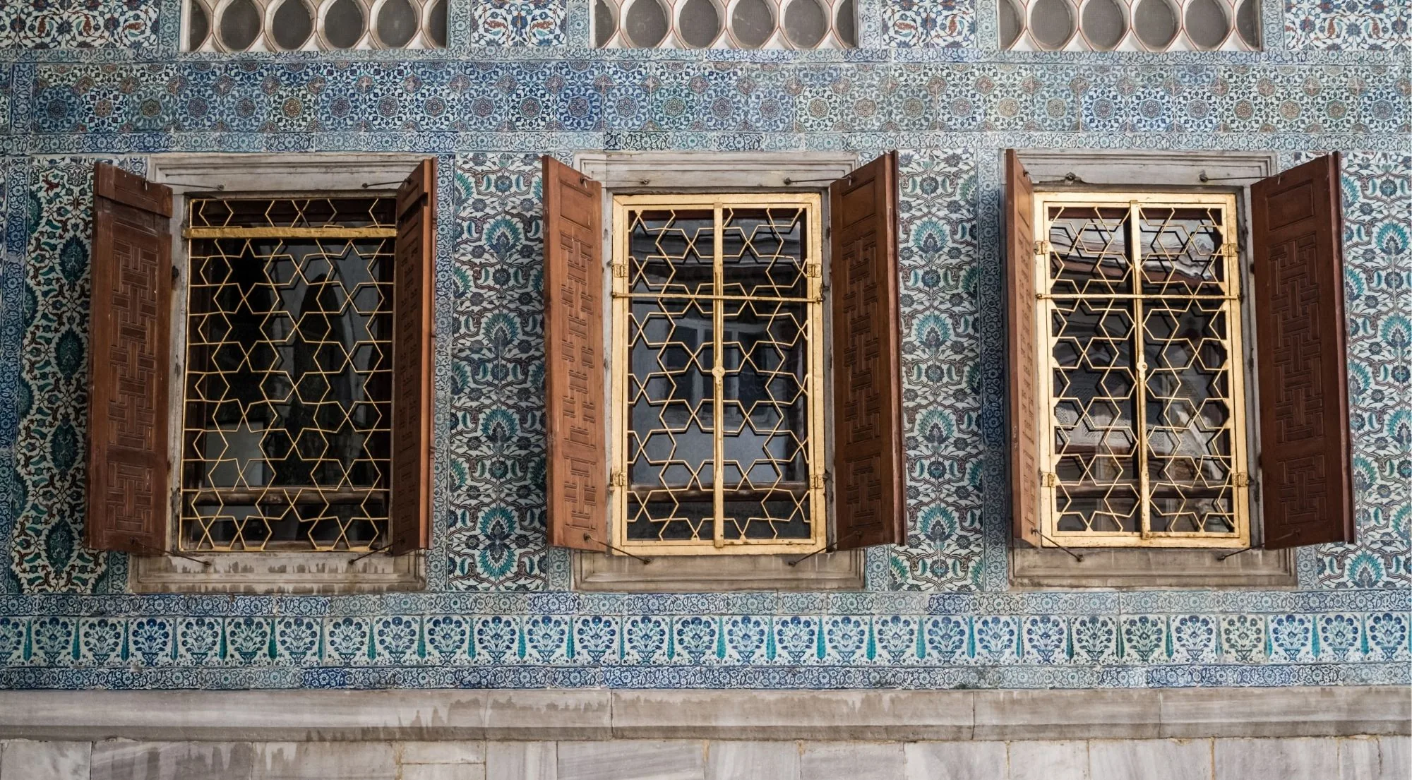 Three shuttered windows with gilded geometric grilles set into a wall of blue and turquoise Iznik tile panels at Topkapi Palace
