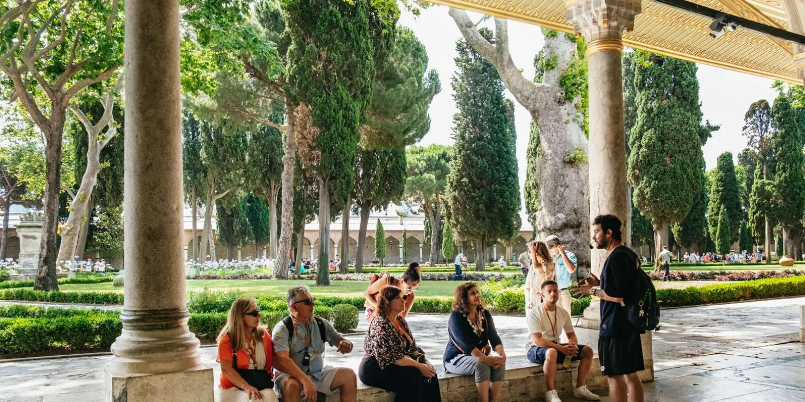 A tour group seated in the shade of a colonnade in a Topkapi Palace courtyard while their guide talks