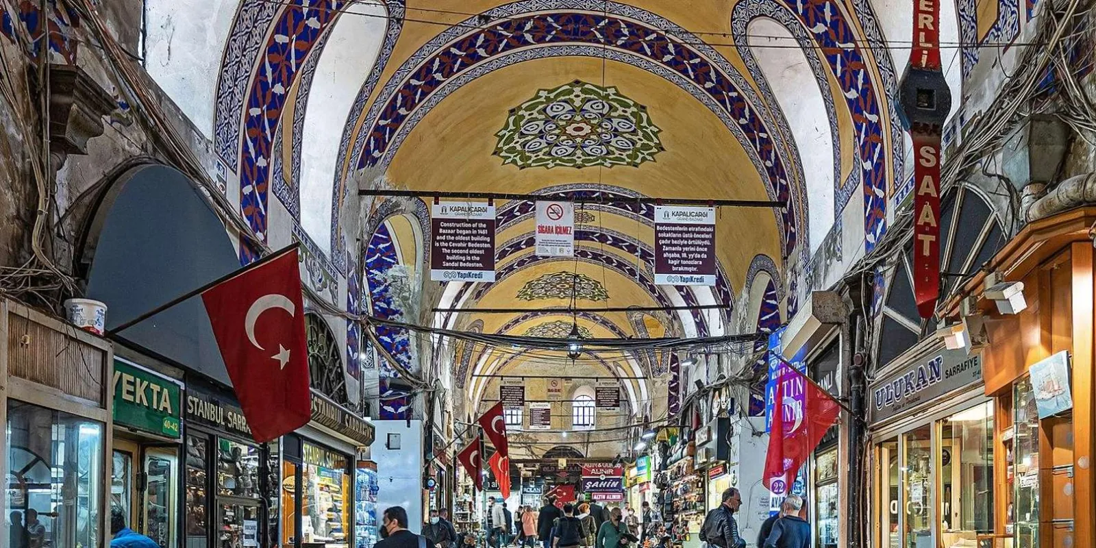 The painted vaulted ceiling and shopfronts of a main lane in the Grand Bazaar, hung with Turkish flags